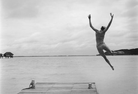 FRIDAY: A woman in a swimsuit jumps off a wooden dock into calm, tropical waters. The image is in black and white and captures the woman in mid-air, with her arms outstretched and legs extended. The background features a calm, still water surface, a distant shoreline, and a few overwater bungalows.