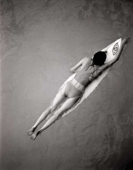 PADDLING: A black and white photograph of a woman lying on a surfboard in shallow water. She is wearing a bikini and has her arms outstretched. The water is calm and the surface is smooth.