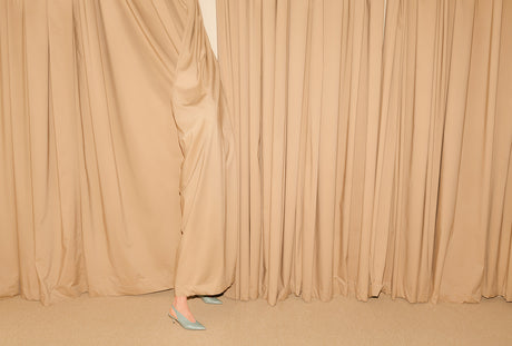 CURTAIN 2: This image shows a womans legs and blue shoes peeking out from behind beige curtains in a room. The curtains are drawn shut, creating a sense of mystery and intrigue. The womans legs are partially visible, adding an element of sensuality to the composition. The image is minimalist and contemporary, with a focus on shape and form.