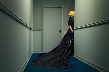 LOSTROOM 2: A woman with bright yellow hair is wearing a long black dress. She stands in a hallway with green walls and a blue carpet, partially obscured by a wall. There is a closed white door to her left, and a portion of another wall to her right.