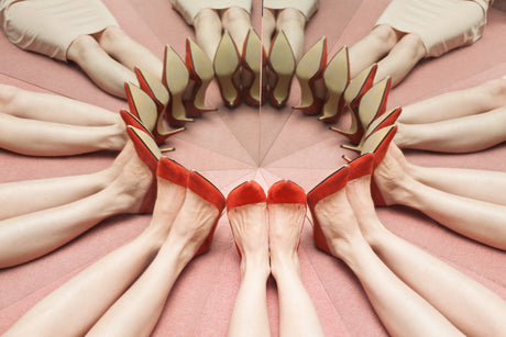 PERFECT STRANGERS: A photograph of several womens legs, all wearing red heels, lying on a pink surface and pointing their toes inwards to create a circular formation. The image is mirrored, creating a symmetrical pattern of legs and heels.