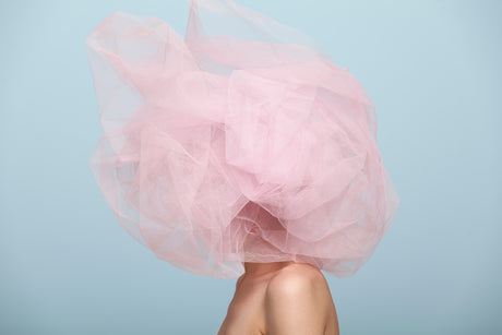 PINK CLOUD: A woman stands against a light blue backdrop with a large amount of pink tulle covering her head and upper body. The tulle is wispy and translucent, creating a soft, dreamy effect. The womans face is obscured by the tulle, leaving only her shoulders and neck visible. The image is a study in color and texture, with the bright pink tulle contrasting sharply with the cool blue background.