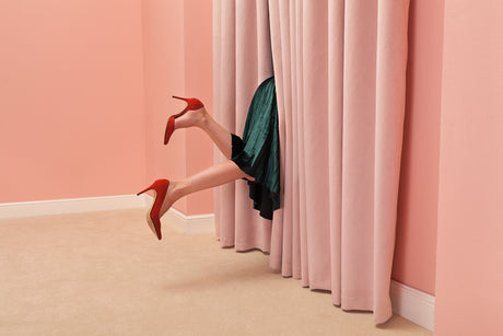 PINK ROOM 2: A womans legs in red high heels are peeking through a pink curtain in a room with pink walls. She is wearing a green skirt and is partially hidden behind the curtain. The photo is taken from a low angle and creates a playful and mysterious atmosphere.