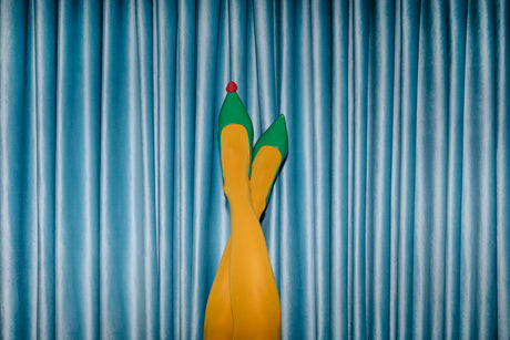 RASPBERRY: A pair of yellow tights with green shoes on the feet are pictured against a backdrop of blue curtains. The feet are pointed up in the air, with the toes of the shoes pointing towards the camera.