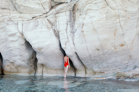 SAZLICA: A woman wearing a red swimsuit stands in a shallow cave opening, facing away from the camera. The cave is carved into a white cliff face, and the woman is standing in the clear, turquoise water.