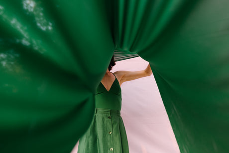 UNTITLED GREEN: A woman is holding up a large piece of green fabric, creating a tunnel-like effect. The fabric is translucent, revealing a glimpse of the womans face and body. The background is a plain white surface.