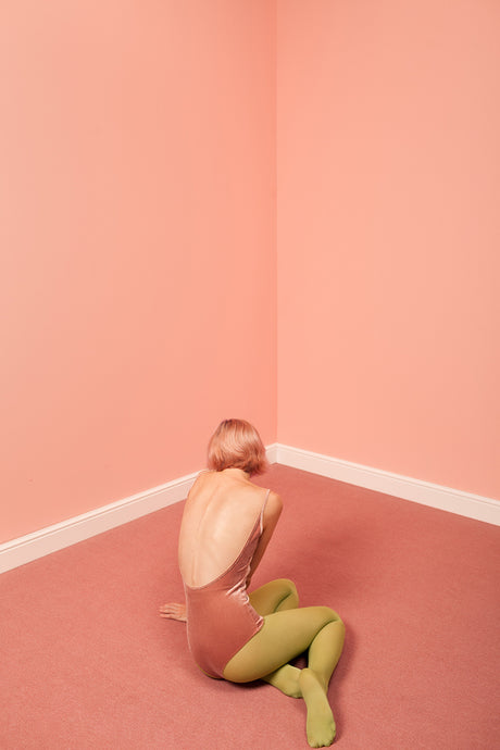 VELVET: A woman with short, blonde hair sits on the carpeted floor in the corner of a room with pink walls. She is wearing a pink bodysuit and lime green tights. She is facing away from the camera and looking down. The floor is a darker pink than the walls.