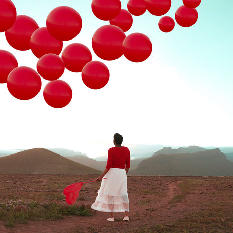 ENDLESS OPTIONS: A woman wearing a red sweater and white skirt stands on a mountain top, gazing upwards at a cluster of red balloons floating in the clear sky above. The mountain landscape is characterized by brown hills and green vegetation, with a distant body of water visible. The woman holds a red net in her right hand, adding a touch of whimsy to the scene.