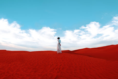 TO A BRIGHT FUTURE: A woman in a white dress stands on top of a red sand dune under a clear blue sky with fluffy white clouds. She is facing away from the camera, looking out at the vast desert landscape.