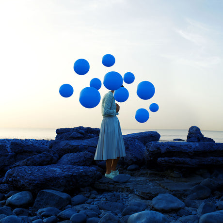 WINTER BLUES: A woman in a white dress stands on a rocky beach at sunset, surrounded by blue balloons that appear to be floating above her head. The balloons are arranged in a cluster, creating a visually striking contrast against the sky and the womans silhouette.