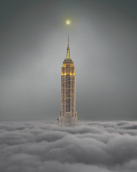 EMPIRE STATE: The Empire State Building rises above a sea of clouds, its golden lights illuminating the twilight sky. The sun sets in the distance, casting a warm glow over the iconic skyscraper.