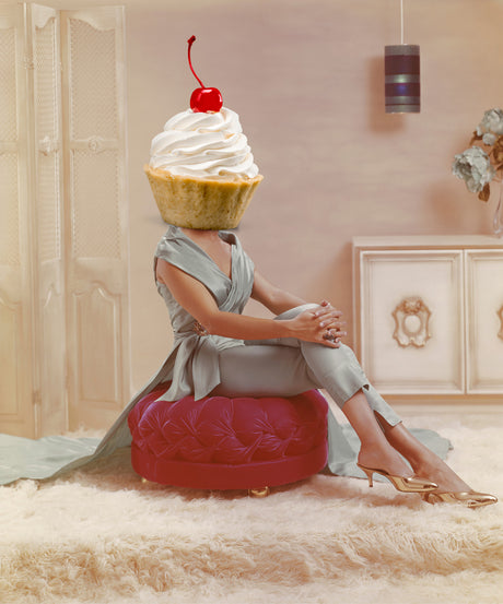 PENANCE: A woman wearing a light blue dress sits on a red velvet ottoman with her legs crossed. She is holding her right hand on her knee and looking off to the side. Her head is replaced with a cupcake topped with whipped cream and a cherry. The photo has a mid-century modern aesthetic, with a white shag rug, a white cabinet in the background, and a simple light fixture. The woman is casually dressed and appears to be relaxed and comfortable.