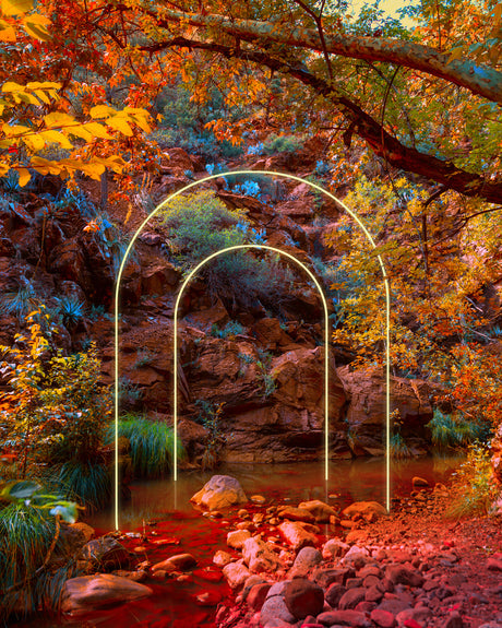 TWO DIMENSIONAL: A photograph capturing the serene beauty of a creek nestled within an autumnal forest. The creek is illuminated by the warm glow of the setting sun, creating a striking contrast with the cool, deep red hues of the rocks and water. Two golden arches, reminiscent of a portal, stand tall against the backdrop of lush greenery and towering trees, inviting viewers to explore the hidden wonders of this magical place.