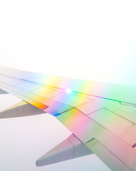 ENTERING RAINBOW: This image shows a close-up of an airplane wing during flight, with rainbow light reflecting off of the surface. The wing is angled down towards the ground, and the light is shining brightly on the surface.