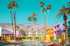 PALM SPRINGS COLORFUL 1: A colorful hotel in Palm Springs, California, features a swimming pool surrounded by lounge chairs and palm trees. The hotel building is a vibrant mix of colors, and the mountains in the background offer a scenic backdrop.