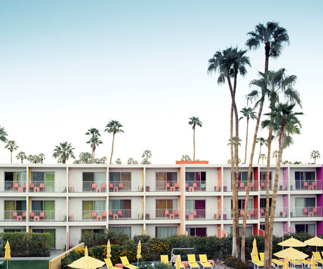 PALM SPRINGS HOTEL: This image showcases a vibrant, colorful mid-century modern motel building in Palm Springs, California. Palm trees line the building, which features bright pink, yellow, and orange accents, and a swimming pool. Lounge chairs are set up around the pool, inviting relaxation and enjoyment of the sunny desert climate.