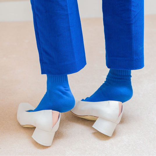 MIISTA: A close-up image shows a person wearing white heels with blue socks. They are wearing blue pants that are folded up above the heel, revealing a portion of the blue socks. The image is captured from an above angle, focusing on the feet and lower legs.