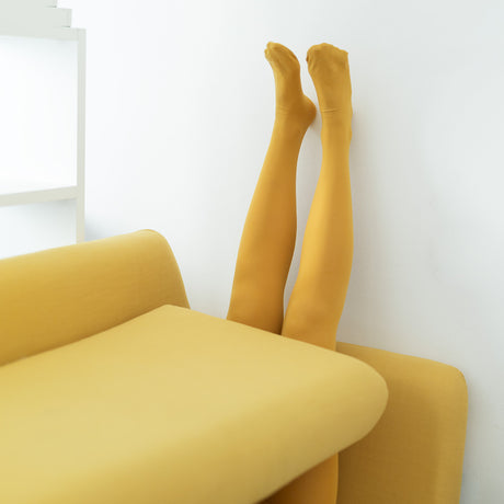 YELLHOW: A pair of yellow tights are visible with the feet pointing towards the ceiling, upside down, and resting against a white wall. The legs are partially hidden by a yellow couch in a room with white walls.