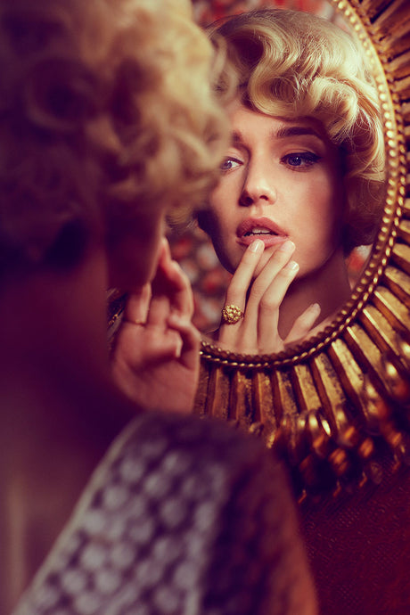 MARILYN 2: A woman with blonde hair and bright makeup looks intensely at her reflection in a large, ornate mirror. The mirror has a golden frame and is decorated with intricate details. The womans hand rests on her chin, and she appears to be admiring her appearance.