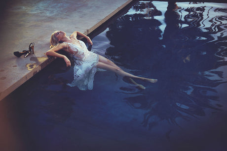 REFLECTED: A woman in a white dress relaxes on the edge of a pool at dusk. Her feet are submerged in the water, and she is looking up at the sky. The pool is dark blue, and the sky is a soft orange.