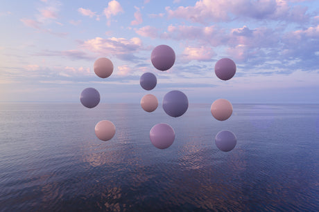 COLLECTIVE: This image shows a cluster of purple and pink balloons floating against a backdrop of a blue ocean and a pastel pink and purple sunset sky. There are puffy white clouds scattered across the sky, and the water below is calm and reflective.