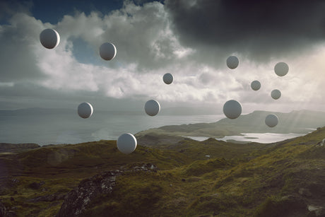 DRIFT: A digital artwork featuring a collection of white spheres floating in the air above a scenic Scottish landscape. The spheres are scattered across the sky, with some appearing closer to the viewer and others farther away. The landscape below is characterized by rolling green hills, a shimmering body of water, and a distant mountain range. The overall atmosphere is serene and dreamlike.