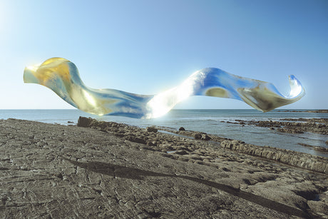 FLEODRADH III: A large, abstract sculpture, resembling a flowing sheet of iridescent material, hovers above a rocky coastline. The sculpture is positioned against a backdrop of blue sky and ocean, with the sun shining brightly. The rocky terrain in the foreground is covered in a pattern of cracks and crevices.