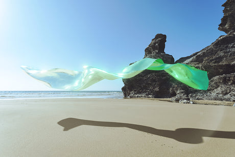 FLEODRADH II: A vibrant green fabric flows gracefully through the air, draping over rugged coastal cliffs. The fabric appears to be translucent, allowing the bright blue sky and the distant ocean to shine through. The sand beach is bathed in sunlight, creating a serene and picturesque scene.