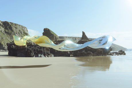 FLEODRADH I: A large, abstract, metallic sculpture is positioned on a sandy beach. The sculpture is made of two pieces, one a gold color and the other a silver color, that flow and curve in a wave-like motion. The sculpture is set against a backdrop of rugged cliffs. The water is calm and reflecting the blue sky.