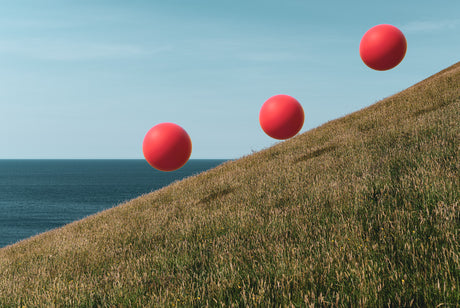 MANSANDS: A still life image featuring three red balloons rolling down a grassy hillside. The hillside slopes down toward a blue ocean, and the sky is a clear blue. The red balloons appear to be rolling downhill, with the first balloon at the top of the hill and the last balloon at the bottom of the hill. The photo is taken from a low angle, giving the viewer a perspective of looking up at the balloons as they roll down the hill.