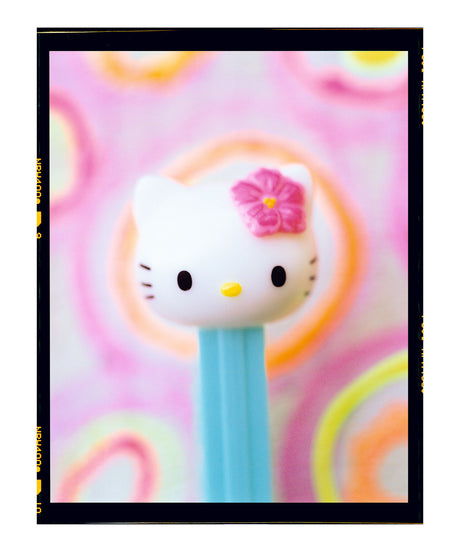 ROXY: A close-up shot of a Hello Kitty Pez dispenser with a pink bow against a brightly colored background. The dispenser is in focus, while the background is slightly blurred.