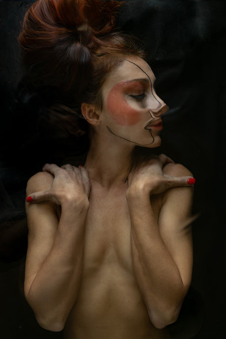 AQVA BLACK: A woman with red hair and bold, theatrical makeup poses for a studio portrait. Her arms are crossed over her chest, and her eyes are closed. The image is shot in a dark, intimate setting with a focus on her face and torso.