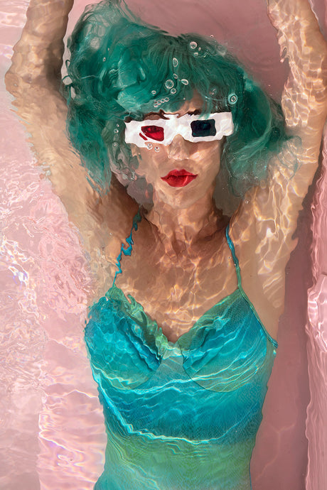 AQVA BLUE: This photo shows a woman with green hair wearing 3D glasses while submerged in a pink bathtub. The water is clear and allows you to see her clearly, as well as the pink surface of the bathtub. The woman is looking directly at the camera.