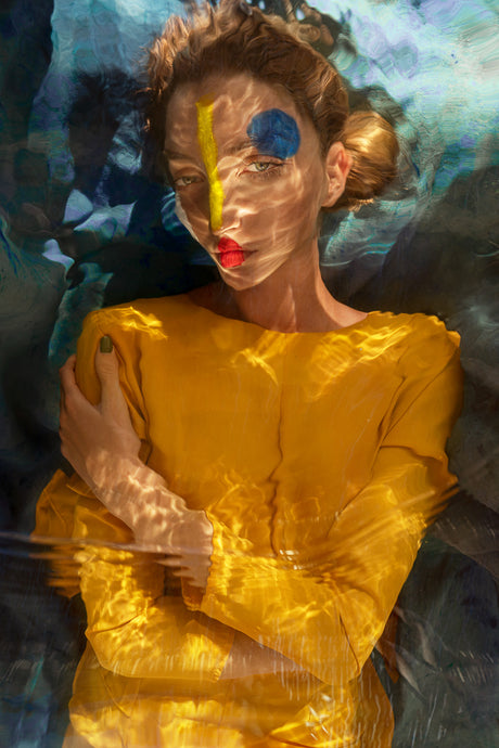 AQVA YELLOW: A woman wearing a yellow dress with painted facial features is submerged in water. The water creates a distorted, dreamlike effect, highlighting the colors and the womans features. Sunlight illuminates the scene, casting shimmering reflections on the water.