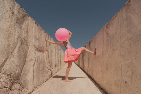 BIG BUBBLE 1: A woman wearing a pink skirt and a silver top is holding a large pink balloon over her head as she stands in between two tall concrete walls. The bright blue sky can be seen above the walls.