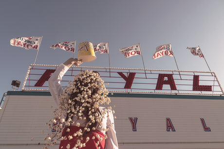 CIRCUS: A woman stands in front of a building with a sign that reads Circus Royal with several flags of the same design in the background. She throws popcorn in the air.