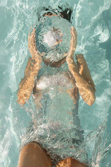 LIGHT UP I: A woman is submerged in a blue pool, holding a disco ball in front of her face with both hands. The water creates a rippling effect around her, and the light reflects off the disco ball.