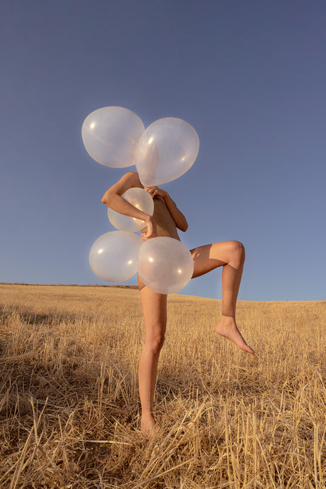 NUDE: A woman is standing in a field of dry grass, holding a bunch of white balloons in front of her. Her body is partially obscured by the balloons, and she is looking up at the sky. The sun is shining brightly, and the sky is a clear blue.