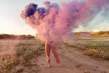 PINK SMOKE: A person stands barefoot in a field with a large cloud of pink smoke billowing around them. The sun is setting behind the smoke, casting a warm glow on the scene. The field is covered in white flowers, and the sky is a pale blue.