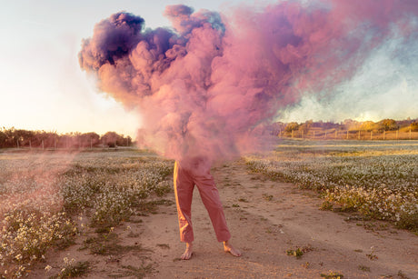 PINK SMOKE: A person stands barefoot in a field with a large cloud of pink smoke billowing around them. The sun is setting behind the smoke, casting a warm glow on the scene. The field is covered in white flowers, and the sky is a pale blue.