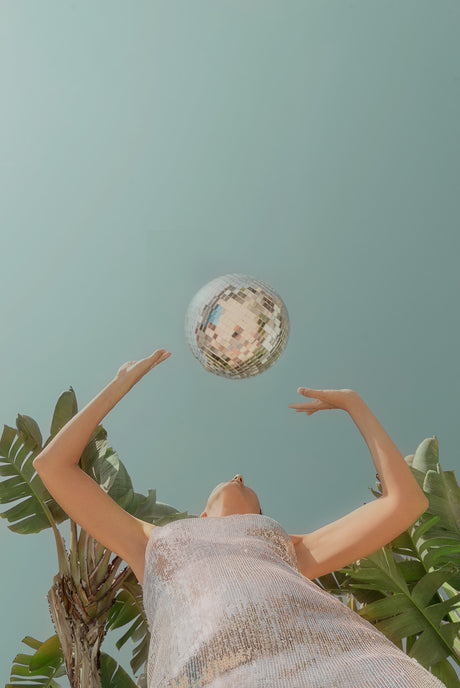 SHINING SHINING: A woman in a silver sequined dress reaches up to catch a disco ball with her outstretched arms. The image is shot from a low angle, and the sky is a light blue, giving the image a dreamy, ethereal quality. Palm trees frame the scene, adding to the tropical feel.