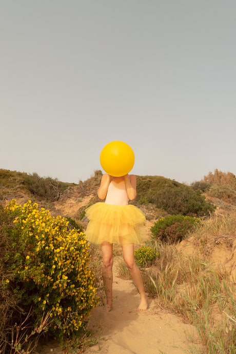 SUNSET DREAM II: A woman wearing a yellow tulle skirt stands on a sandy path in a beach landscape. She is holding a large, yellow balloon in front of her face, covering her eyes. The sun is shining brightly in the sky, creating a warm, golden hue over the scene. The womans bare feet are visible on the sand as she walks through the brush.