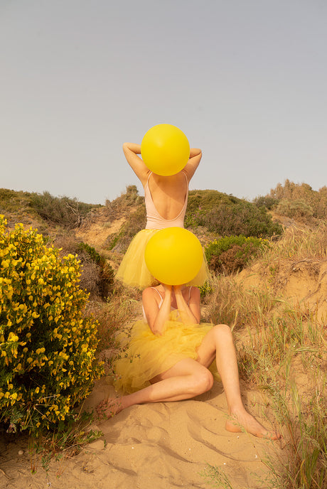 SUNSET DREAM: Two women playing with yellow balloons in a sandy, outdoor setting. One woman stands in a yellow tulle skirt, holding a balloon over her head, while the other sits in the sand, using a balloon to cover her face. The scene evokes a sense of playful whimsy and carefree joy, creating a cheerful and lighthearted atmosphere.