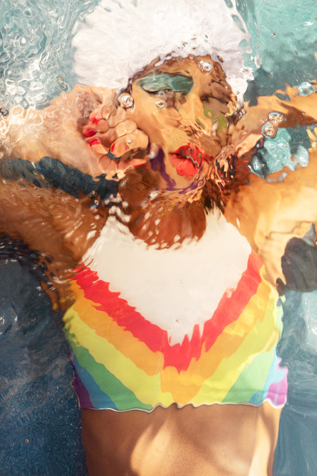 THE SHAPE OF WATER III: A woman with a white swim cap and a rainbow swimsuit is underwater. The water is blue and the womans face is visible through the water. She appears to be holding her breath.