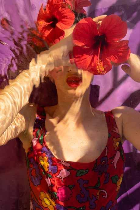 THE SHAPE OF WATER II: A woman is submerged in a pool, her face partially obscured by vibrant red hibiscus flowers. She wears a red floral swimsuit and her hair is dark and wet. Sunlight shines through the water, illuminating her face and the flowers, creating a dramatic and elegant scene.