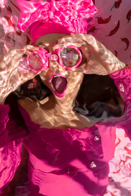 THE SHAPE OF WATER IV: This image features a woman wearing pink sunglasses while submerged underwater. She is wearing a pink shirt and a pink headband, and there are red peppers scattered throughout the background. The image is taken from a slightly above angle, giving a view of the womans open mouth and surprised expression.