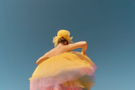 YOU ARE MY RAINBOW II: The image shows a woman wearing a yellow tulle dress with a pink tulle underskirt, and a yellow tulle bow in her hair. She is facing away from the camera, with her back to the viewer. She is holding the skirt with one hand, and her other arm is extended. The background is a bright blue sky. The photo has a dreamy and whimsical quality.