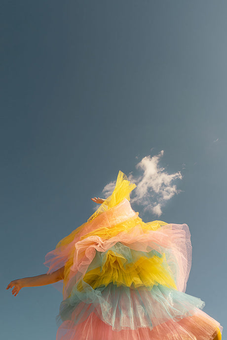 YOU ARE MY RAINBOW: A woman wearing a multi-layered, pink, yellow, and blue tulle tutu jumps in the air against a backdrop of a bright blue sky. The skirt billows around her as she leaps, and her arms are outstretched. The sun shines brightly, casting a warm glow on the scene.