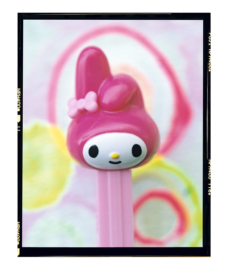 SASSY: A close-up photograph of a pink Hello Kitty Pez dispenser in focus against a blurred background of pastel pink, yellow, and green circular shapes. The Hello Kitty characters iconic pink ears and bow are prominently displayed. The photo has a soft, dreamy aesthetic.