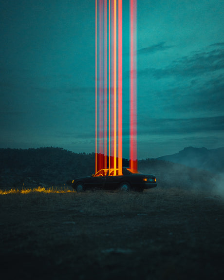 ABDUCTION: The image depicts a black car parked on a grassy field at dusk, illuminated by a vibrant, multicolored light show in the sky. The light streaks appear to be caused by long-exposure photography, capturing the movement of light sources. The dark sky is a rich teal blue, and the surrounding landscape is hazy and muted, adding to the ethereal atmosphere of the scene.